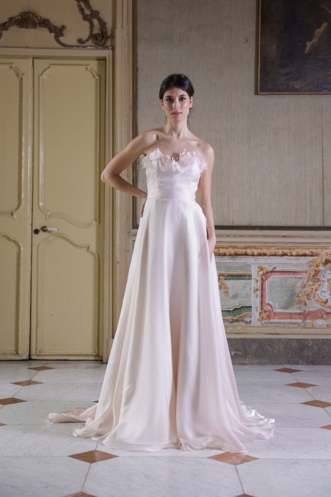In questa foto, una sposa indossa un abito da sposa scivolato color cipria chiaro, con corpino senza spalline decorato da petali delicati tono su tono che impreziosiscono il décolleté. La gonna, morbida e leggera, scivola elegante fino a terra, creando un look romantico e senza tempo. La scena è ambientata in una sala storica, con pareti affrescate e porte antiche.