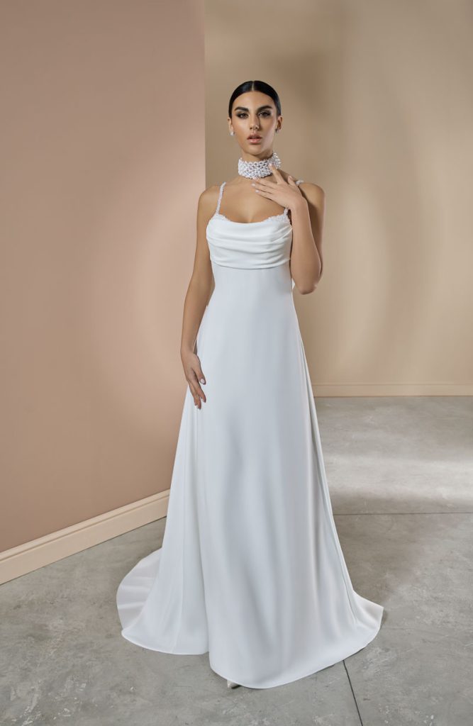 In questa foto, una modella indossa un abito da sposa scivolato con taglio dritto e tessuto fluido. Il corpino ha una scollatura drappeggiata e sottili spalline gioiello, mentre un raffinato choker di perle aggiunge un tocco moderno e sofisticato. L’ambiente neutro e minimalista valorizza la pulizia delle linee dell’abito.