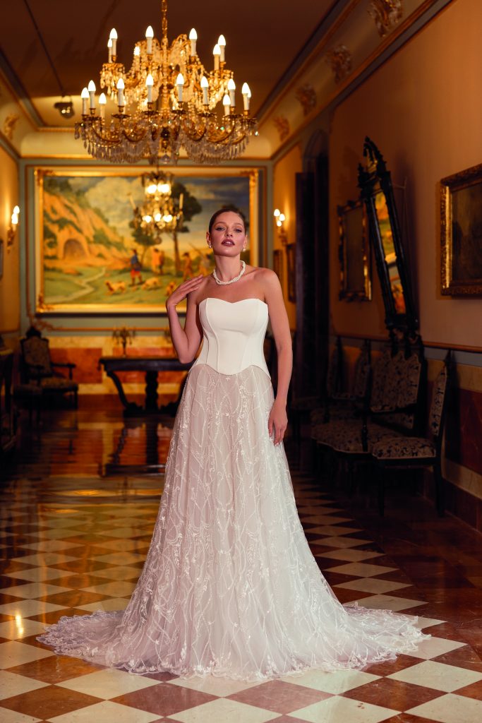 In questa foto, una sposa indossa un abito da sposa scivolato 2026 con corpetto strutturato a cuore e una gonna ampia in tulle ricamato. Il contrasto tra il bustino liscio e la leggerezza trasparente della parte inferiore crea un effetto raffinato ed etereo. La scena si svolge in una lussuosa sala con pavimento a scacchi e sontuosi lampadari dorati.