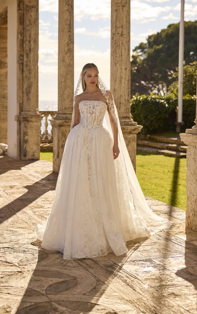 In questa foto, una modella posa all’aperto sotto un portico in pietra, indossando un sontuoso abito da sposa scivolato 2026 in pizzo. Il corpetto, strutturato e semitrasparente, è decorato con ricami floreali, mentre il velo lungo e ricamato completa il look con un tocco classico. Sullo sfondo, si intravedono il mare, un giardino verde e la luce calda del tramonto.