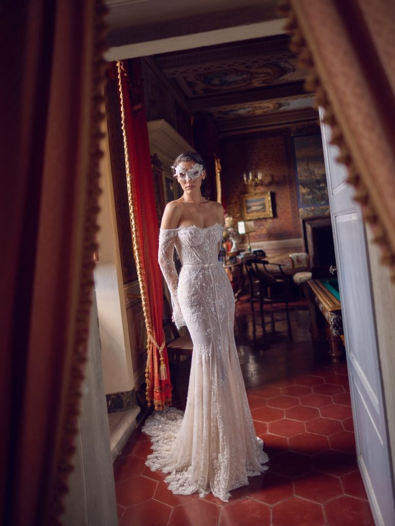 In questa foto, una modella posa in un sontuoso ambiente d’epoca, con un abito avvolgente e riccamente decorato di pizzi e perline. Le maniche scese e lo strascico fluente aggiungono teatralità e sensualità alla linea a colonna tipica degli abiti da sposa scivolati.