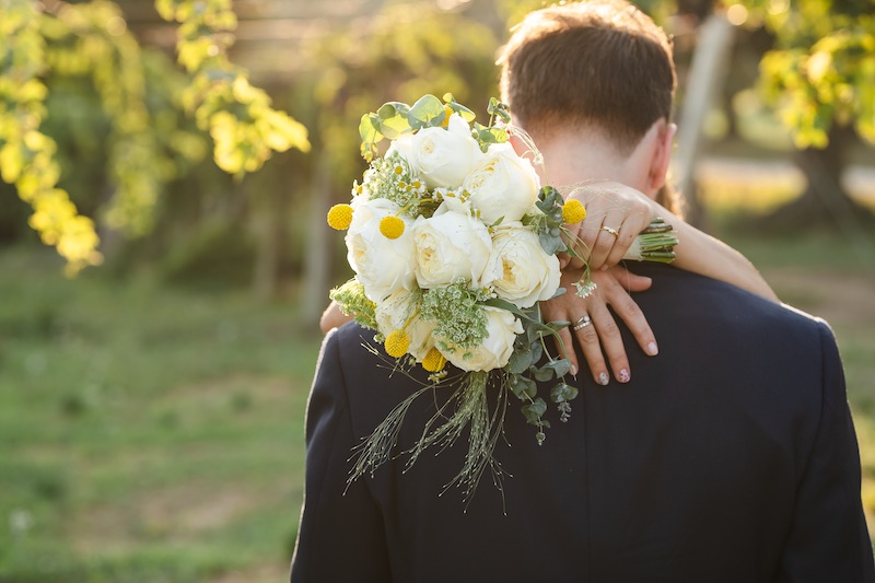 In questa foto, due sposi abbracciati di spalle: è inquadrato il dettaglio del bouquet di rose inglesi di colore bianco e piccoli fiori gialli tenuto in mano dalla sposa nella mano sinistra