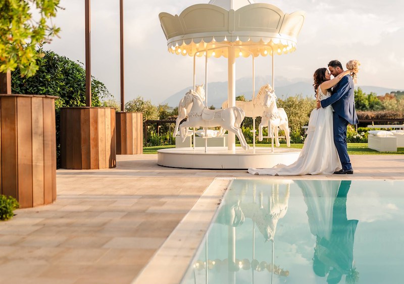 In questa foto, due sposi di un matrimonio organizzato da Ada Di Donato: si trovano a bordo piscina sul quale si riflettono gli sposi abbracciati davanti ad un Carousel di colore bianco