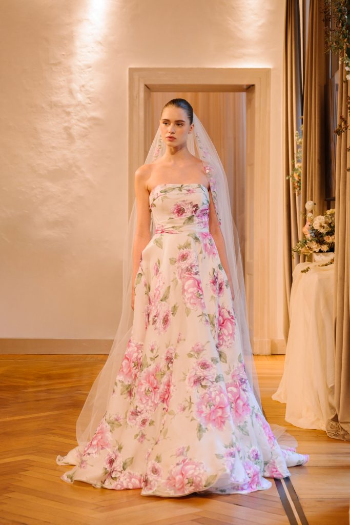 In questa foto, una modella indossa un abito da sposa con stampa floreale rosa e verde su fondo avorio. Il vestito, senza spalline, ha un taglio ampio e romantico ed è abbinato a un lungo velo. Fa parte della collezione abiti da sposa Atelier Emé 2026.