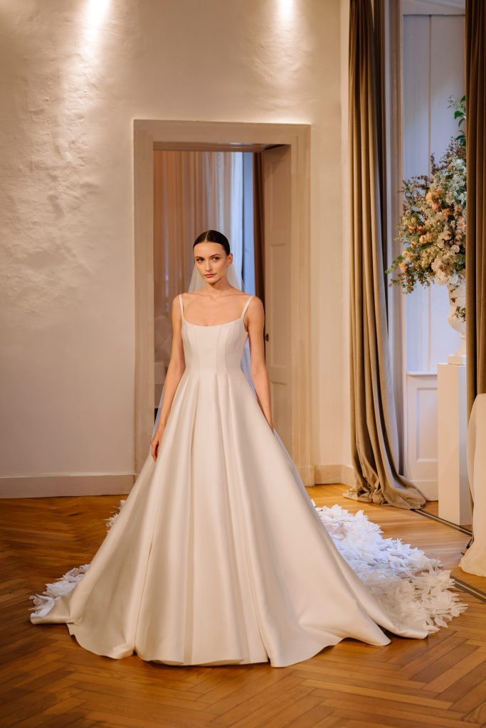 In questa foto, una modella indossa un elegante abito da sposa con spalline sottili e scollo squadrato, caratterizzato da una linea semplice e scivolata in tessuto lucido. La gonna si apre in un'ampia gonna a campana con un lungo strascico decorato da applicazioni floreali in rilievo. La scena è ambientata in un'elegante sala con pavimento in parquet e luci soffuse. Questo abito fa parte della collezione abiti da sposa Atelier Emé 2026.