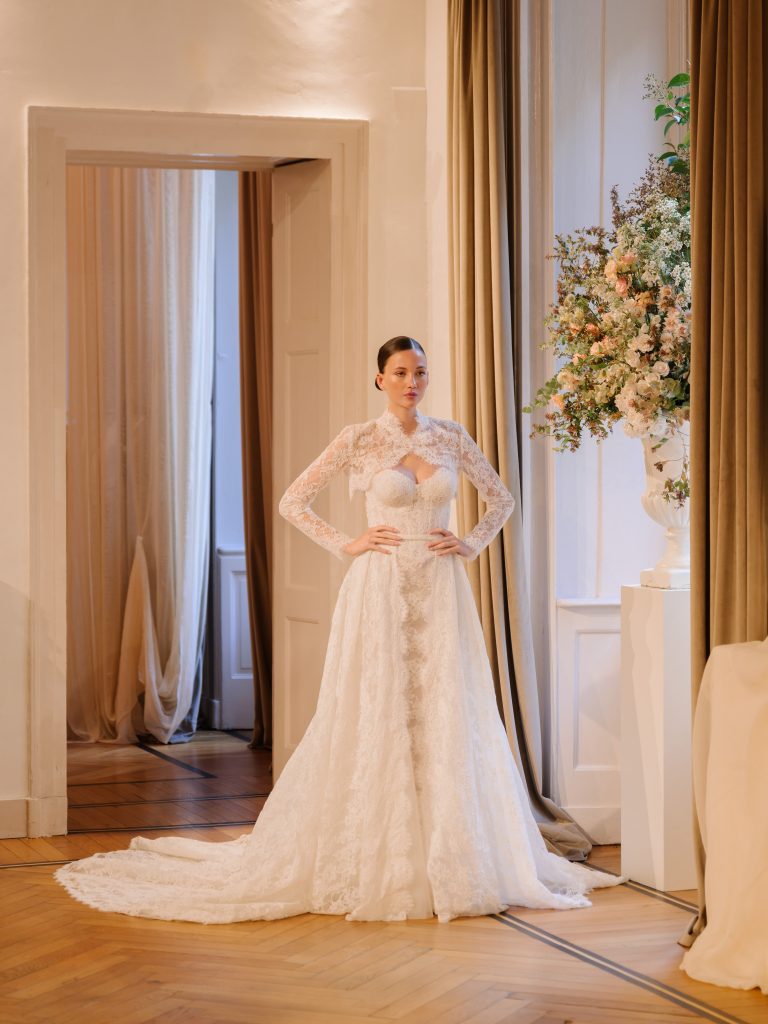 In questa foto, una modella sfoggia un abito da sposa in pizzo con maniche lunghe e corpetto trasparente decorato da raffinati ricami floreali. Il vestito ha un ampio strascico in pizzo che scivola elegantemente sul pavimento. Sullo sfondo, una sala luminosa con tendaggi beige e un grande vaso di fiori completa l’atmosfera sofisticata. L'abito appartiene alla collezione abiti da sposa Atelier Emé 2026.
