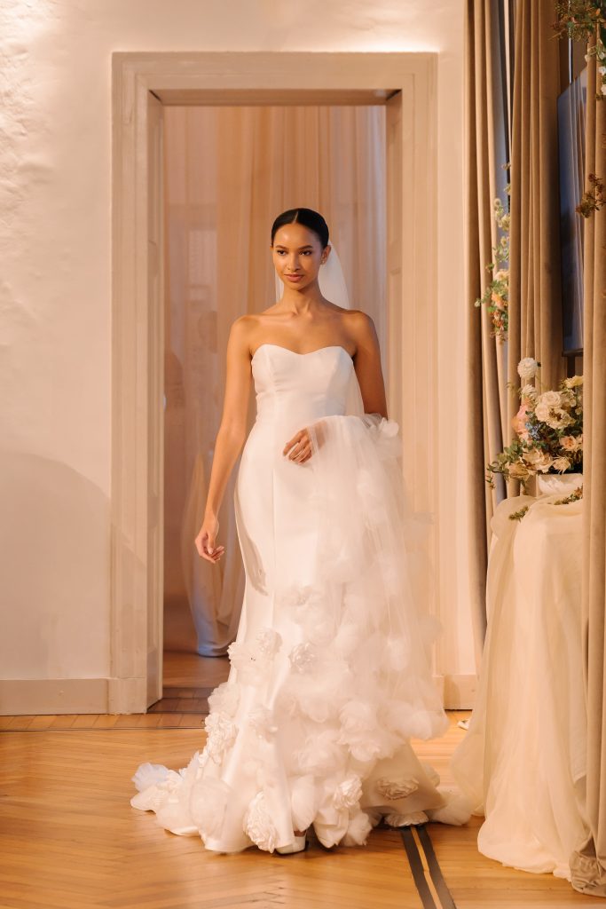 In questa foto, una modella indossa un abito con lungo strascico ricamato e velo coordinato, in un'elegante sala dallo stile classico. Il vestito, parte della collezione abiti da sposa Atelier Emé 2026, è impreziosito da petali in rilievo che decorano il bordo del velo 