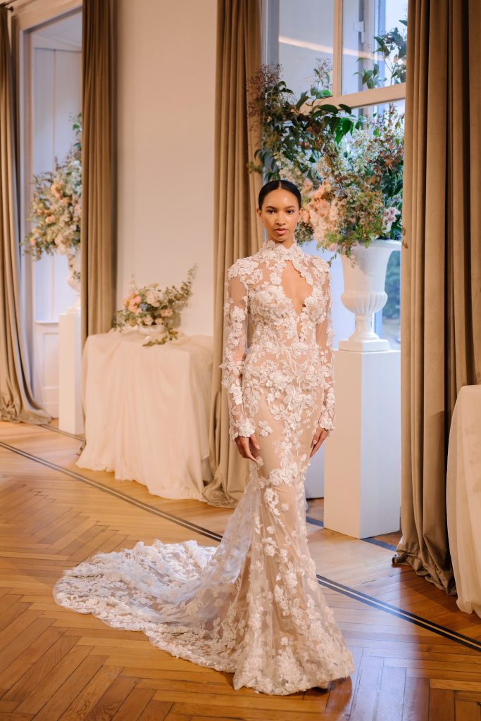 In questa foto, una modella sfila in un raffinato salone con un abito in pizzo trasparente a maniche lunghe della collezione abiti da sposa Atelier Emé 2026. Il modello aderente ha un profondo scollo a goccia e una lunga coda, per un effetto seducente e sofisticato.