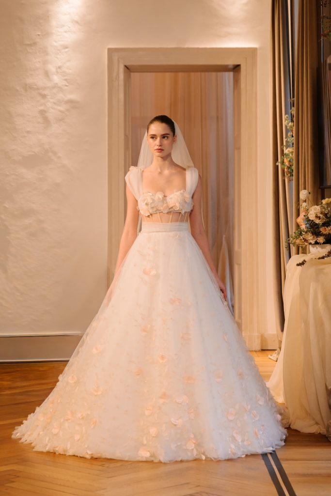 In questa foto, una modella percorre la passerella interna con un abito da sposa principesco della collezione abiti da sposa Atelier Emé 2026. Il vestito ha un corpino decorato con fiori in rilievo tono su tono e una gonna ampia in tulle costellata di petali, mentre il pubblico osserva incantato.