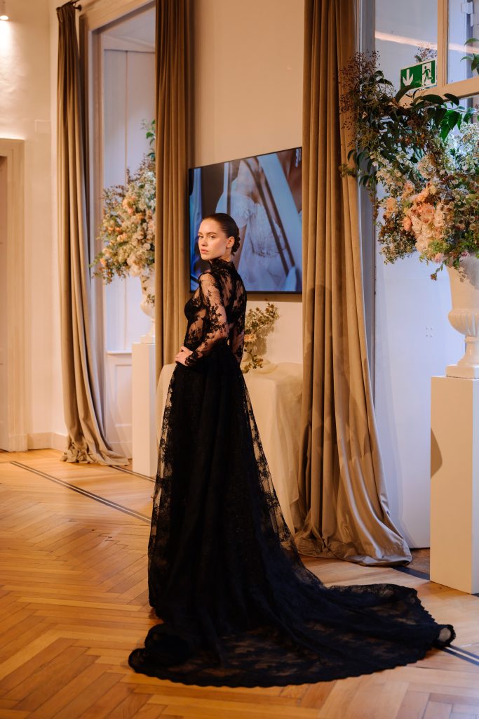 In questa foto, una modella indossa un abito da sposa nero in pizzo con maniche lunghe e collo alto, caratterizzato da una silhouette morbida e una lunga coda. Il vestito fa parte della collezione abiti da sposa Atelier Emé 2026.