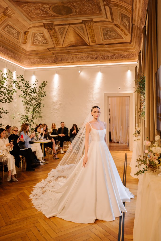 In questa foto, una modella percorre la passerella interna con un abito da sposa principesco della collezione abiti da sposa Atelier Emé 2026. Il vestito ha un corpino decorato con fiori in rilievo tono su tono e una gonna ampia in tulle costellata di petali, mentre il pubblico osserva incantato.