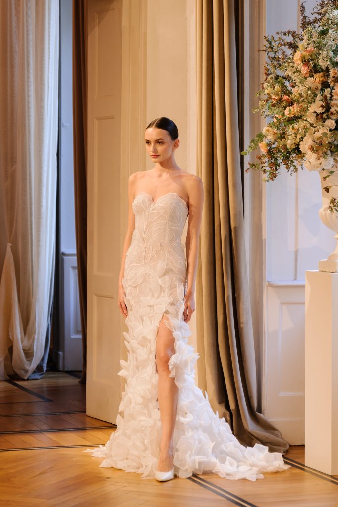 In questa foto, una modella sfila con un abito a sirena in mikado bianco con scollo a cuore, dalla collezione abiti da sposa Atelier Emé 2026. L’orlo è decorato da maxi fiori in tessuto, mentre un velo vaporoso scende dalla sua mano, completando il look con eleganza moderna.