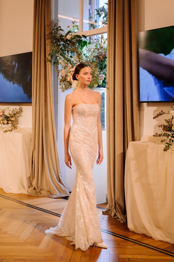 In questa foto, una modella sfila con un abito a sirena senza spalline, interamente ricamato con paillettes e perline. Il tessuto brillante si adagia delicatamente sulle forme, per un effetto elegante e sensuale. L’abito appartiene alla collezione abiti da sposa Atelier Emé 2026.