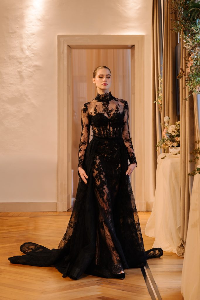 In questa foto, una modella cammina in passerella indossando un abito nero in pizzo trasparente, con maniche lunghe, collo alto e dettagli sartoriali d'effetto. Un design audace e teatrale della collezione abiti da sposa Atelier Emé 2026.