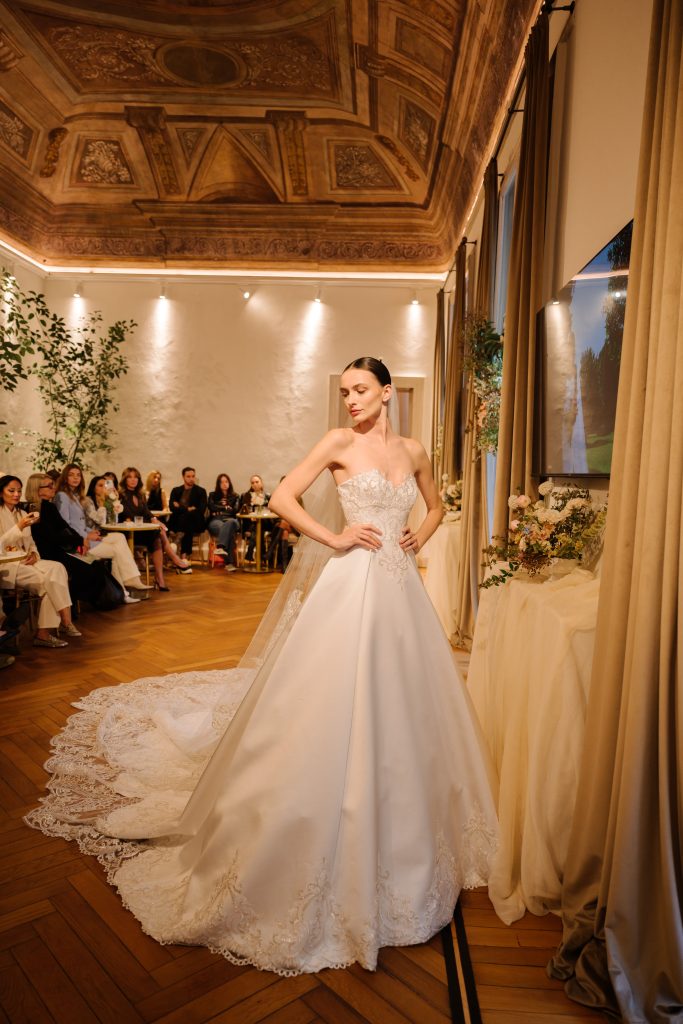 In questa foto, una modella indossa un elegante abito da sposa con ampia gonna e strascico ricamato, mentre sfila in una sala sontuosa con soffitto affrescato e parquet in legno. Il corpetto, senza spalline, è decorato da delicati motivi in pizzo, e il lungo velo completa il look regale. La scena fa parte della collezione abiti da sposa Atelier Emé 2026, presentata di fronte a un pubblico attento seduto lungo i lati della sala.