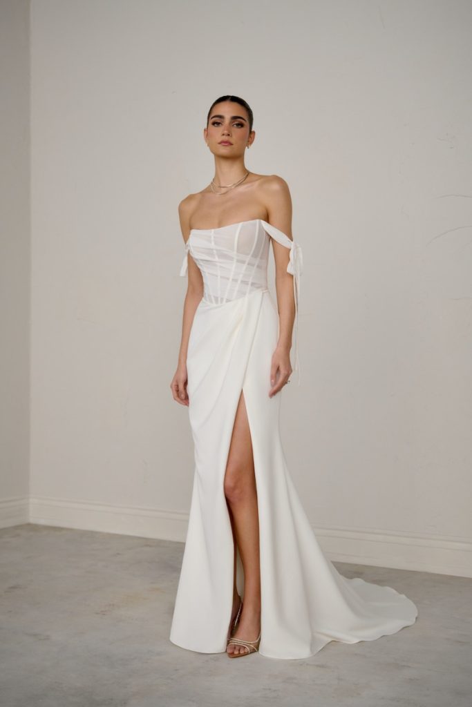In questa foto, una modella posa   indossando un abito a sirena della collezione sposa Justin Alexander Signature P/E 2026 con corpetto steccato trasparente, spacco frontale, fiocchi laterali che scendono lungo le braccia
