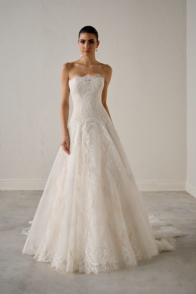 In questa foto, una modella posa  accovacciata indossando un abito ad A della collezione sposa Justin Alexander Signature P/E 2026 modello A-line in pizzo con scollatura dritta 