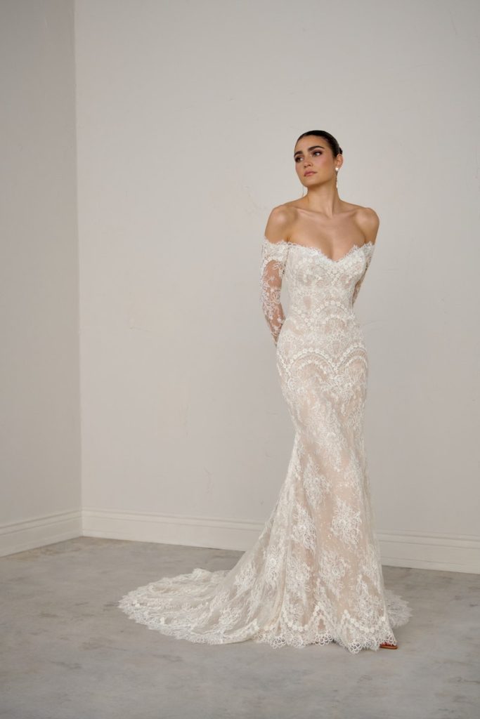 In questa foto, una modella posa indossando un abito a sirena in pizzo della collezione sposa Justin Alexander Signature P/E 2026 con scollo omerale e maniche lunghe