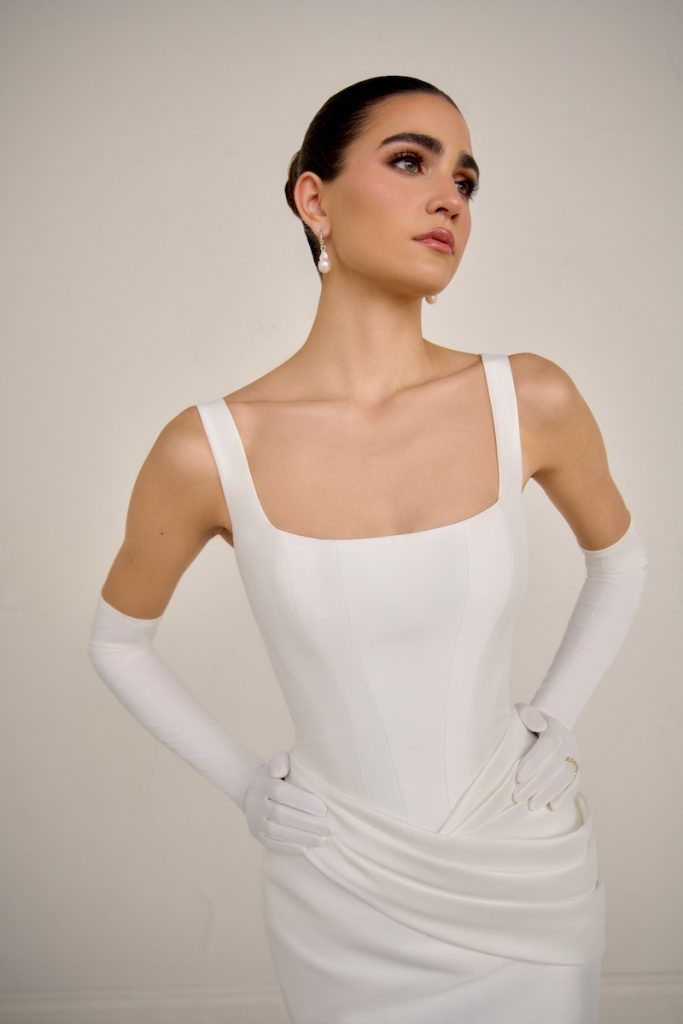 In questa foto, una modella inquadrata a mezzo busto indossando un abito a sirena Justin Alexander Signature con scollo quadrato, drappeggio sui fianchi. Il look è completato con guanti lunghi