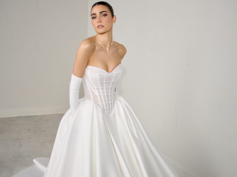In questa foto, una modella posa indossando un abito della collezione sposa Justin Alexander Signature P/E 2026 con corpetto a vita basca trasparente