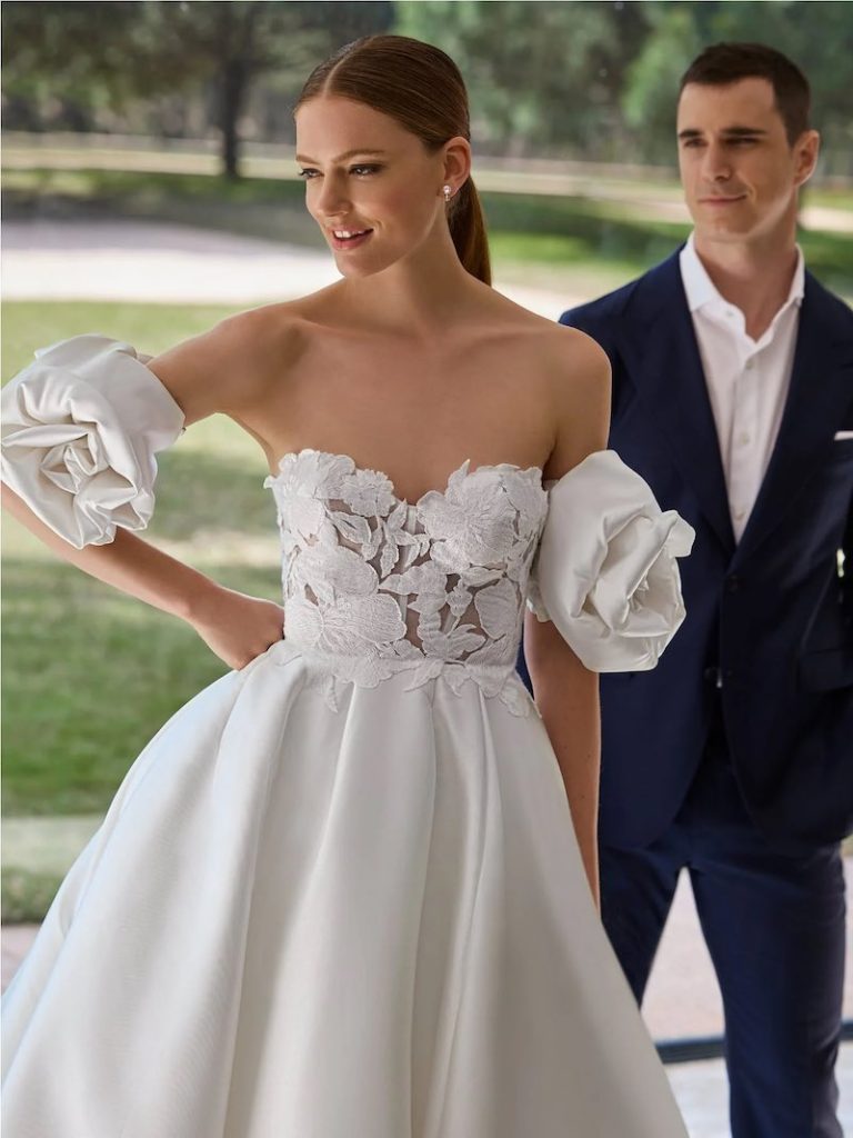 In questa foto, una modella inquadrata a mezzo busto posa con un modello in abito blu elegante indossando un abito da sposa Pronovias 2026 modello ad A, in tessuto strutturato, con corpetto decorato con intarsi floreali e manicotti staccabili a forma di rosa