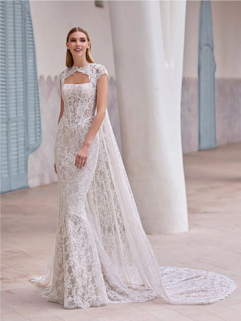 In questa foto, una modella posa indossando un abito da sposa Pronovias 2026 modello a sirena in pizzo Chantilly con cappa abbinata