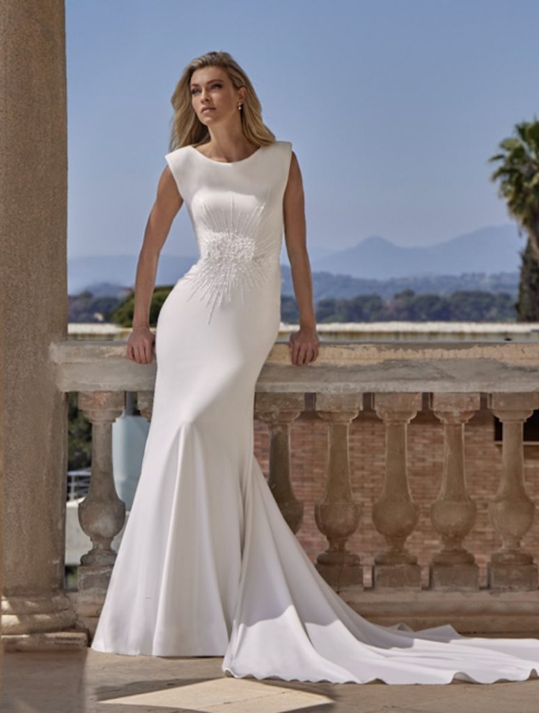 In questa foto, una modella posa indossando un abito da sposa in cady, modello a sirena, con scollo girocollo, ricamo di perline e cristalli a raggiera e lungo strascico