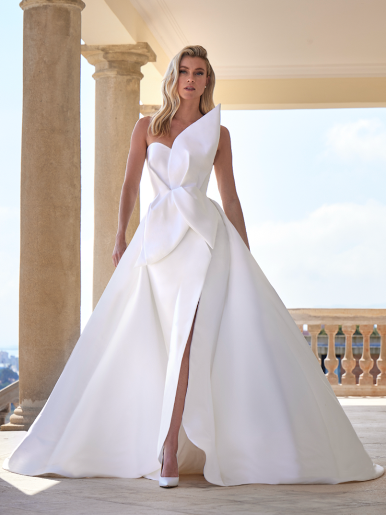 In questa foto, una modella posa indossando un abito della collezione sposa Pronovias 2026 con silhouette  A-Line in tessuto strutturato, con spacco frontale e fiore tridimensionale che si estende sul corpetto fino al punto vita