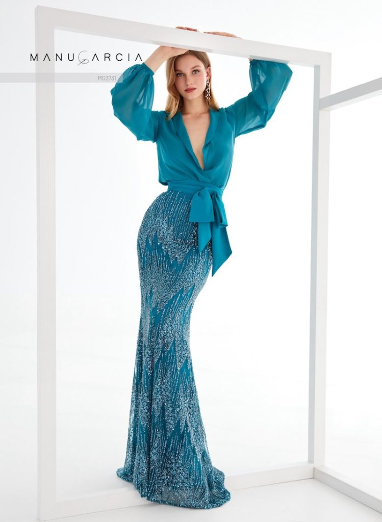 In questa foto, una modella posa indossando un abito da cerimonia Manu García 2026 con silhouette a sirena, gonna ricamata con perline e camicia con scollo a V e fiocco in vita