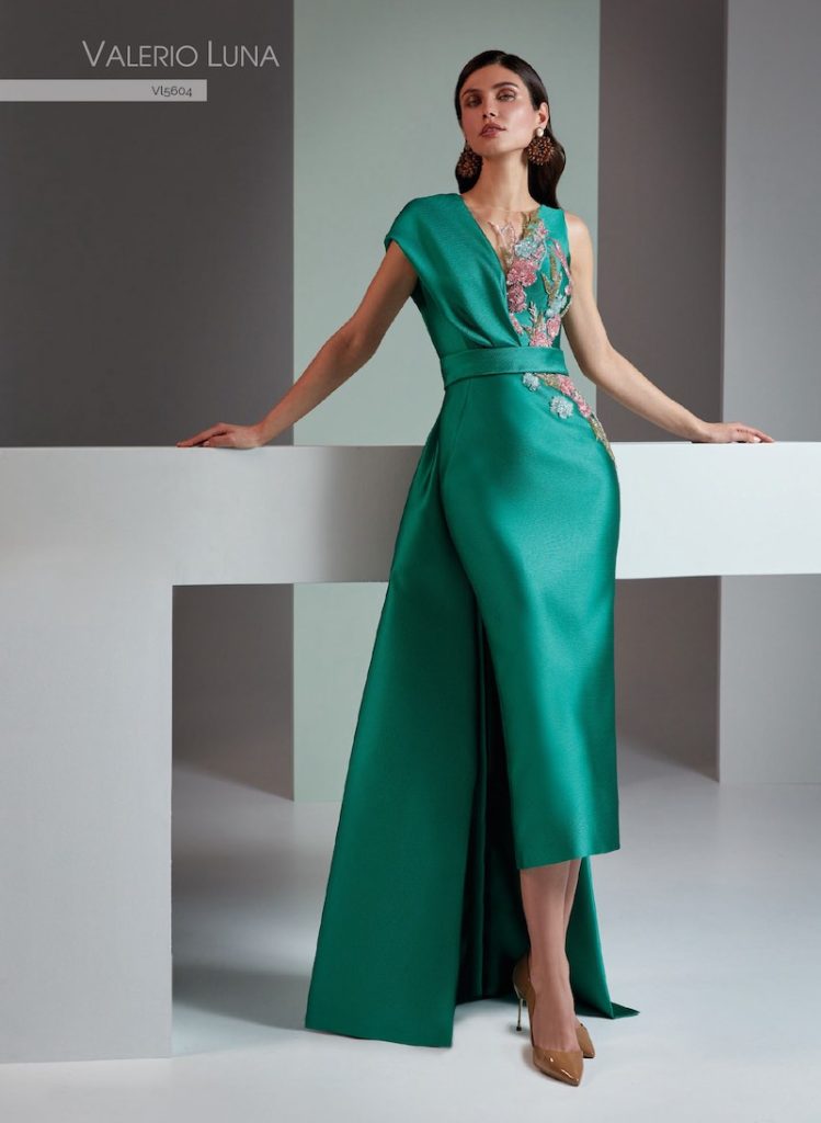 In questa foto, una modella posa indossando uno degli abiti da cerimonia Higar Novias Group 2026 della linea Valerio Luna 2026 modello midi sotto il ginocchio nei toni del verde, con lungo strascico e decorazioni floreali nei toni del rosa