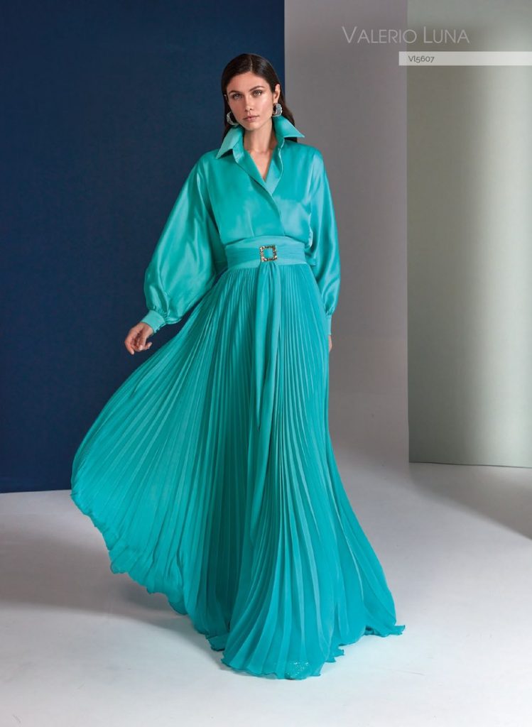 In questa foto, una modella posa indossando uno degli abiti da cerimonia Higar Novias Group 2026 della linea Valerio Luna modello lungo eni toni del verde acqua, con gonna plissé, blusa morbida con maniche a sbuffo, cintura gioiello in vita 