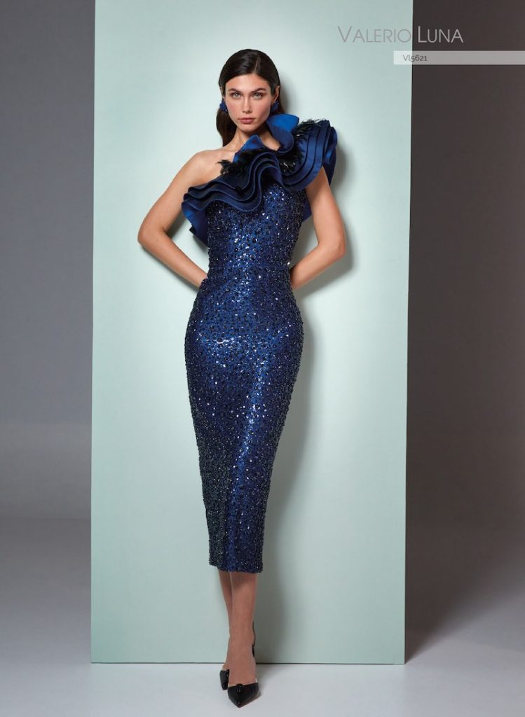 In questa foto, una modella posa indossando un abito midi a sirena con cristalli e ruches lungo la scollatura nei toni del blu della linea Valerio Luna della collezione cerimonia Higar Novias Group 