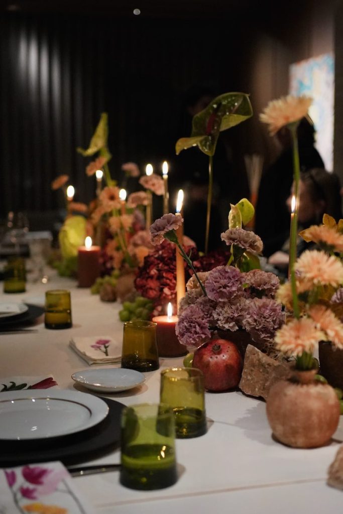In questa foto un tavolo per matrimonio decorato con fiori di colore rosa, pesca e bordeaux, candele, melagrane, bicchieri verdi e tovaglioli dipinti con fiori