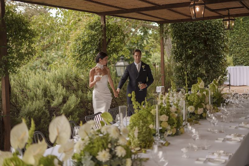 In questa foto due sposi sorridenti che camminano dietro ad un tavolo imperiale decorato per il loro matrimonio da Filomena D'Amelio con fiori di colore bianco e verde