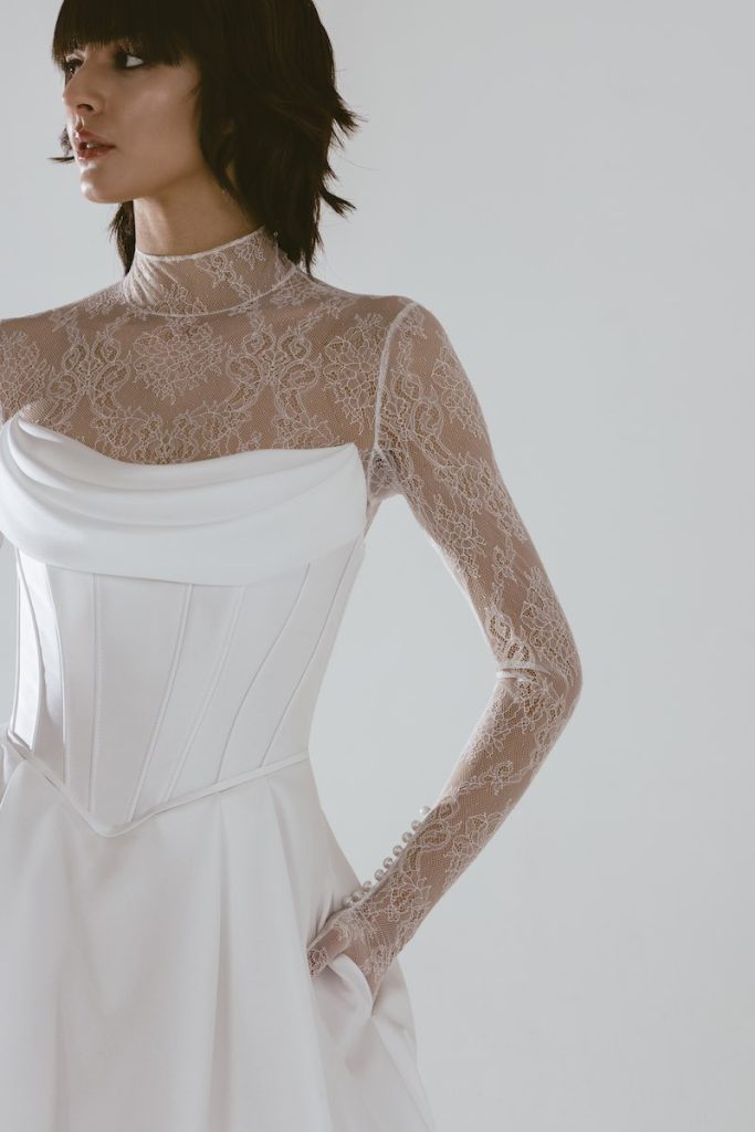 In questa foto una modella inquadrata a mezzo busto posa indossando un abito da sposa con corpetto steccato a vita basca e body in pizzo floreale con maniche a guanto