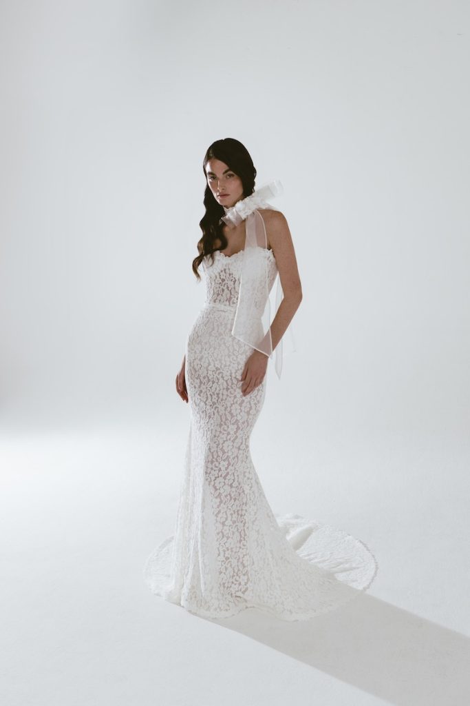 In questa foto una modella posa indossando un abito da sposa a sirena in pizzo floreale della linea Futura by Luisa Lotti, con corpetto con scollo a cuore. La modella indossa anche un chocker maxi con fiocco