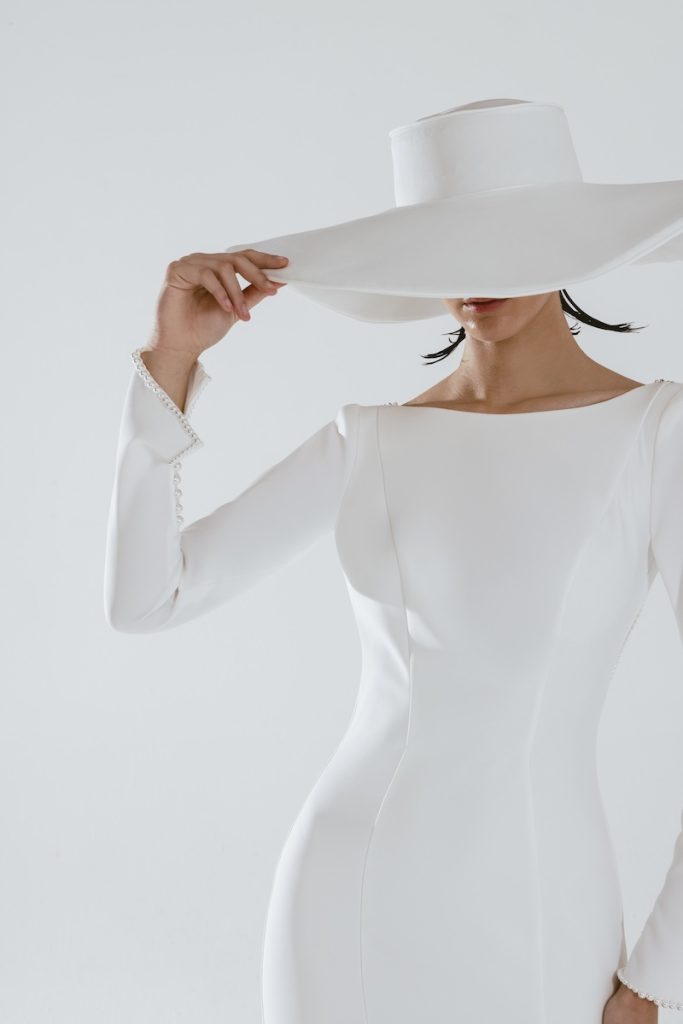 In questa foto una modella posa indossando un abito da sposa a sirena in tessuto stretch con maniche lunghe e scollo a barca della linea Futura by Luisa Lotti. L'abito è abbinato ad un cappello a tesa larga