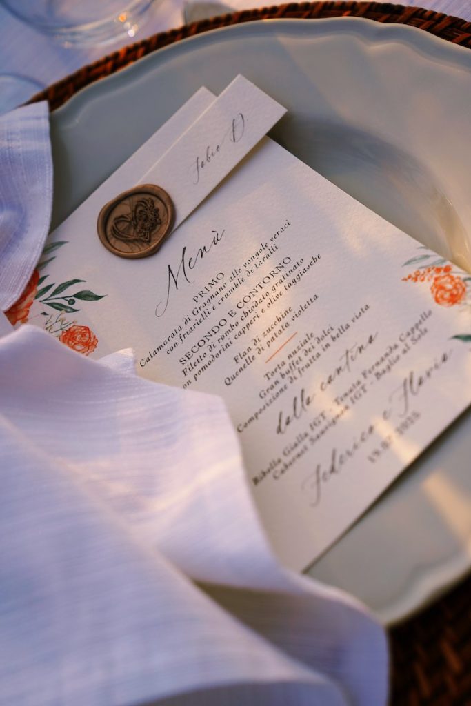 In questa foto il dettaglio di una mise en place del matrimonio di Federico e Flavia realizzata da Manuela Marrale con un piatto bianco, un sottopiatto di rattan, un menù personalizzato  con ceralacca e segnaposto e un tovagliolo bianco annodato