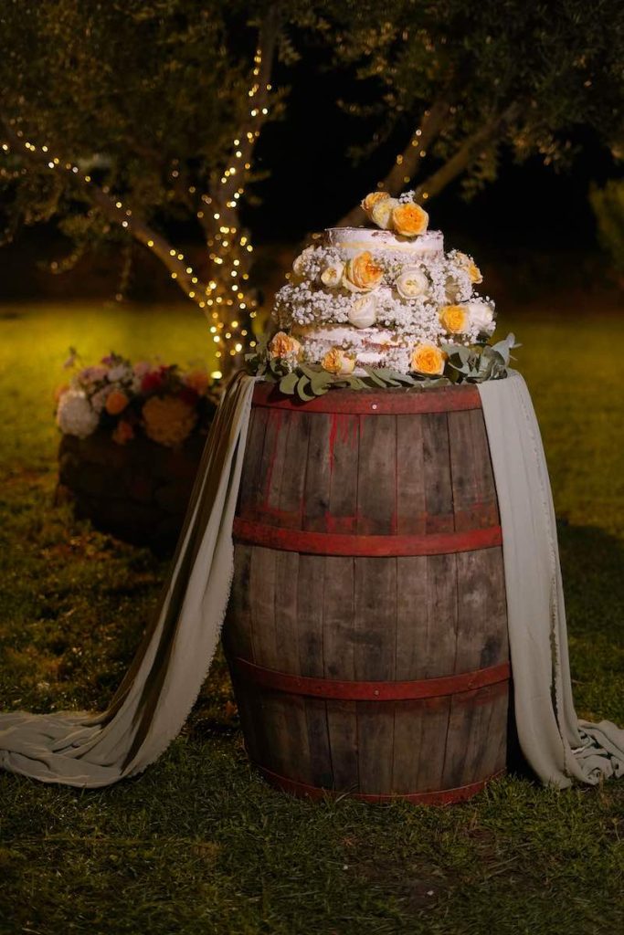 In questa foto, l'allestimento di una torta per matrimonio su una botte di legno decorata con un drappeggio di tessuto verde. La torta è a piani e decorata con fiori di colore giallo e bianco