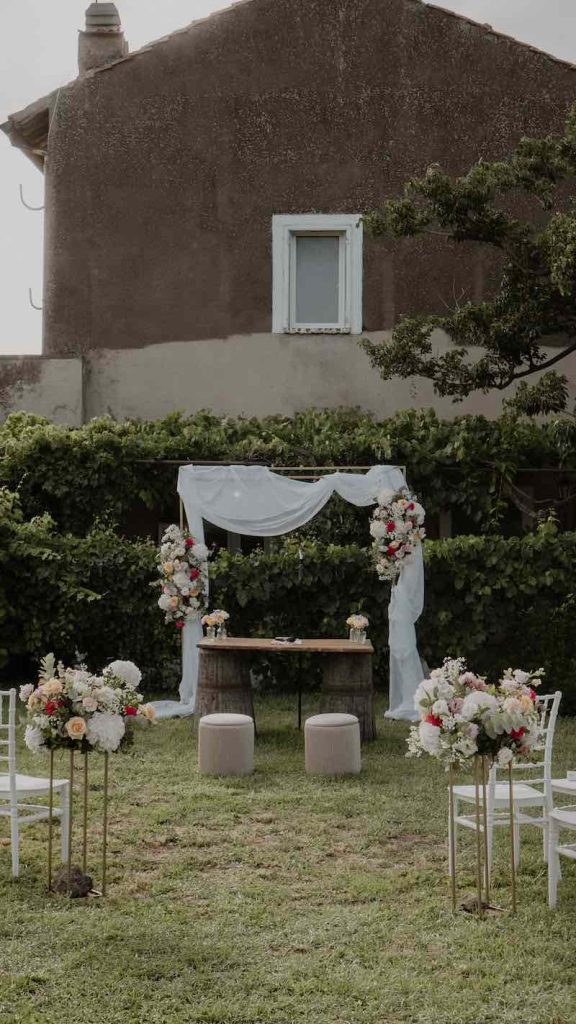 In questa foto l'allestimento di una cerimonia simbolica all'aperto realizzato con un arco con drappeggi di stoffa bianca, fiori colorati, botti e puff di velluto