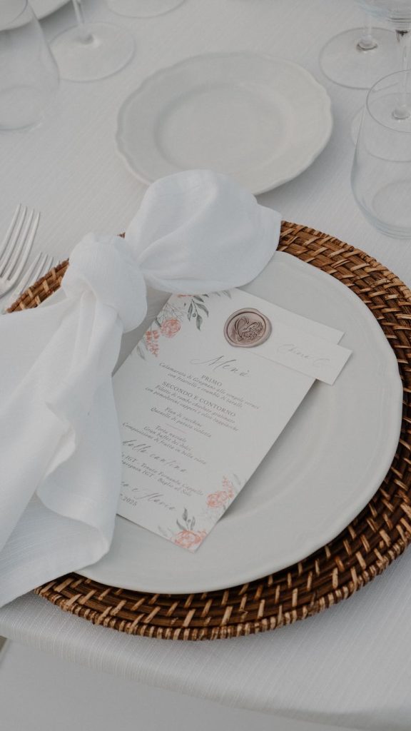 In questa foto il dettaglio di una mise en place del matrimonio di Federico e Flavia realizzata da Manuela Marrale con un piatto bianco, un sottopiatto di rattan, un menù personalizzato  con ceralacca e segnaposto e un tovagliolo bianco annodato