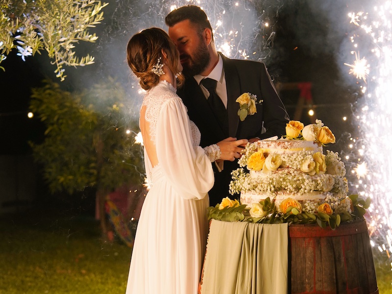 In questa foto due sposi si baciano dietro alla loro torta nuziale decorata con rose di colore bianco e giallo allestita su una botte decorata con un drappeggio verde