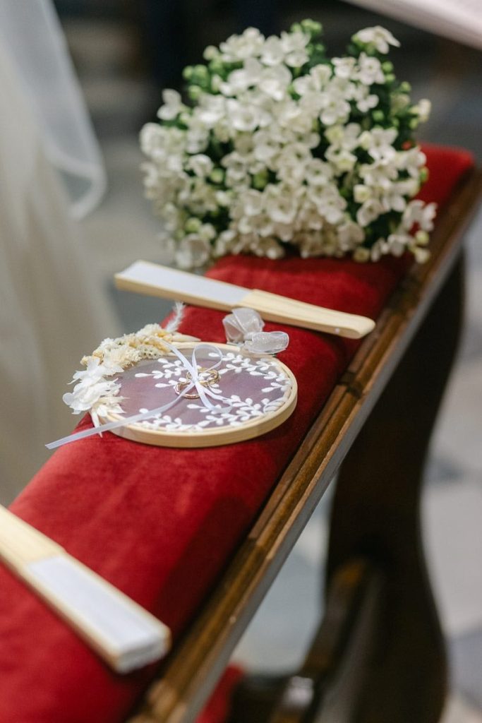 In questa foto un portafedi ricamato su un telaio e poggiato su un inginocchiatoio di velluto di colore rosse, tra due ventagli e un bouquet di mughetto