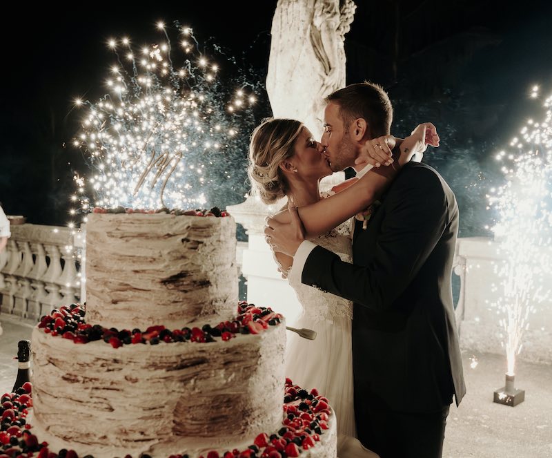 In questa foto, due sposi si baciano dietro ad una torta a piani decorata con frutti di bosco, circondati dallo scoppio di fuochi d'artificio