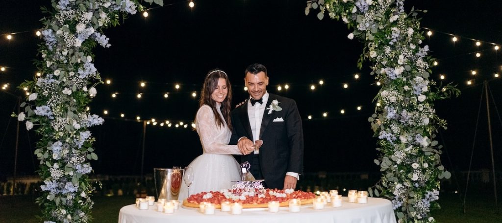 In questa foto, due sposi di un matrimonio firmato dal Wedding Creator Mariano Minetto tagliano una torta nuziale ad un piano davanti ad un arco floreale