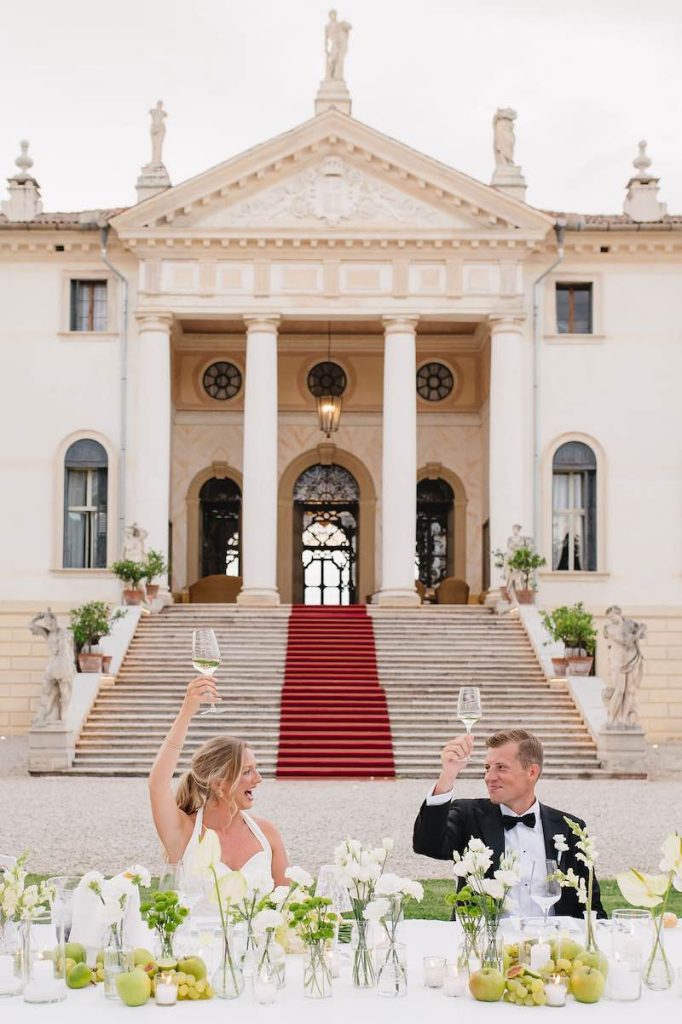In questa foto due sposi brindano seduti ad un tavolo imperiale decorato con mele di colore verde e fiori di colore biancoa davanti alla facciata di una villa storica