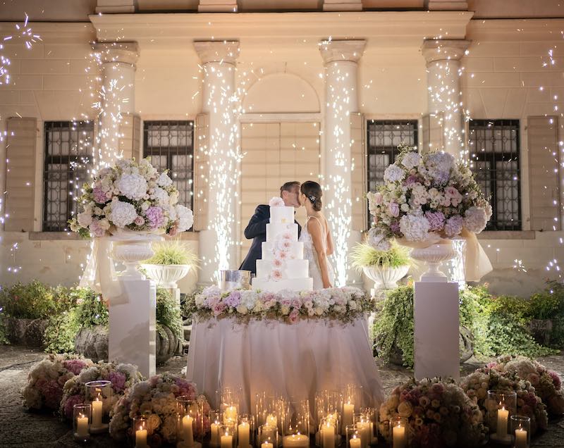 In questa foto, due sposi si baciano dietro ad una torta nuziale a cinque piani decorata con fiori di colore rosa e bianco tra fiori in toni di rosa e fuochi d'artificio