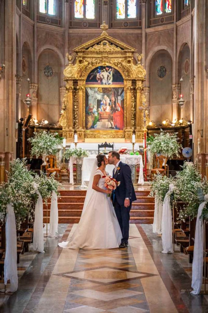In questa foto, due sposi si baciano davanti all'altare di una chiesa decorata con drappeggi di stoffa di colore bianco e fiori abbinati