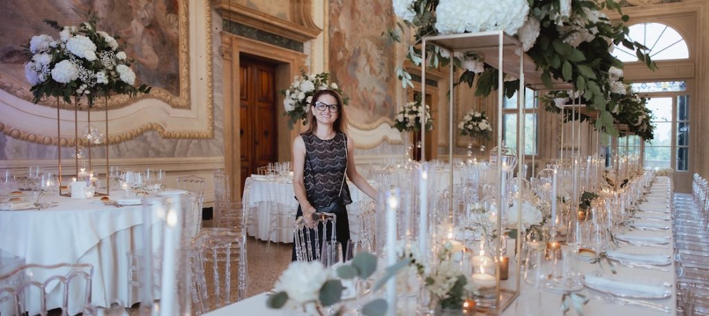 In questa foto, la Wedding Planner Silvia Baldan di Maison Mariage che posa tra tavoli di un matrimonio in una sala elegante allestita con fiori Total White