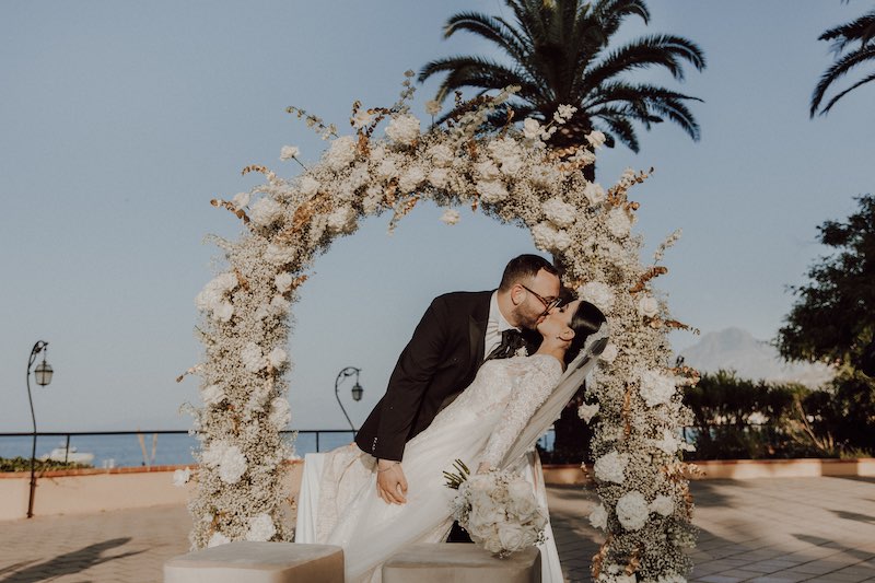 In questa foto, due sposi si baciano davanti all'arco della cerimonia di nozze allestito con gypsophila bianca. Lo sposo fa fare un casquet alla sposa che tiene il bouquet nella mano sinistra